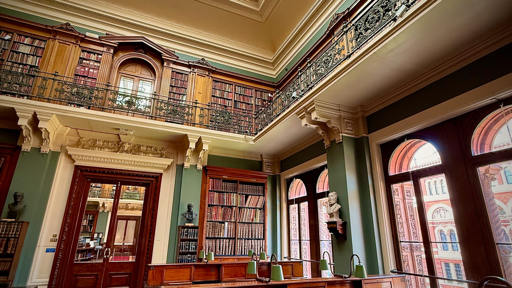 old fashioned library with a vaulted ceiling and mezzanine floor of shelves, dark wood shelving, desks and bankers lamps